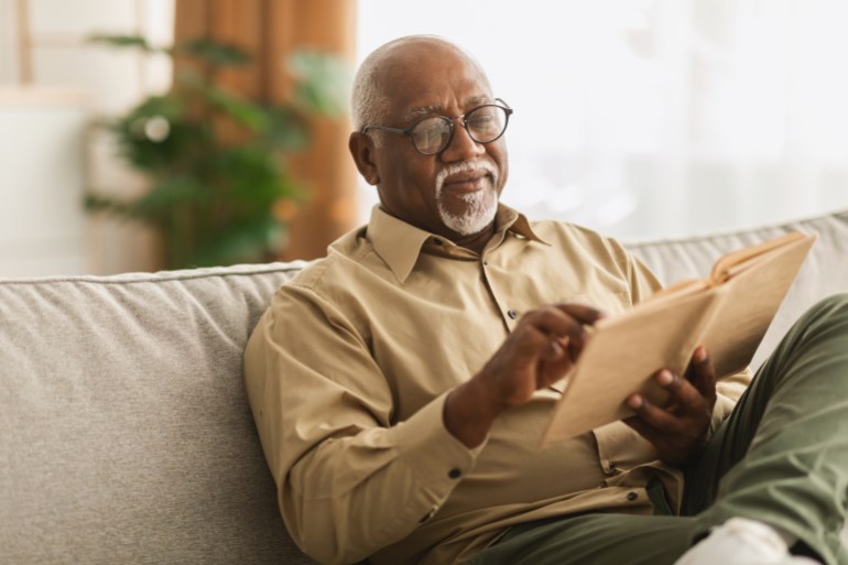 senior african american man reading book sitting on couch at home, wearing eyeglasses. retired male enjoying reading new novel or business literature on weekend. retirement leisure concept; shutterstock id 2079163267; purchase_order: aljazeera ; job: ; client: ; other: