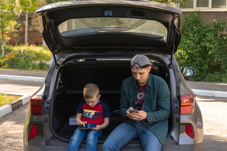 father and son ignore each other using their smartphones. they sit in the trunk of the car and do not pay attention to each other. concept of modern smartphone addiction شترستوك