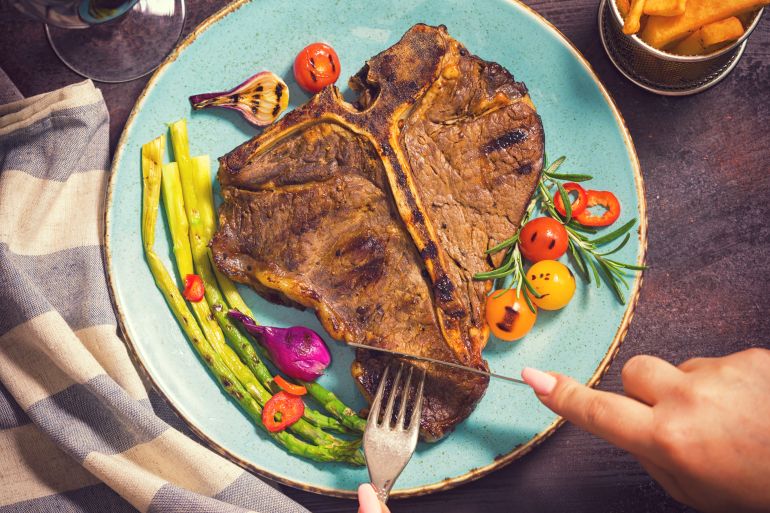 top view of young women eating juicy t-bone steak with french fries,asparagus,roasted cherry tomatoes and red chili peppers over dark rustic background.