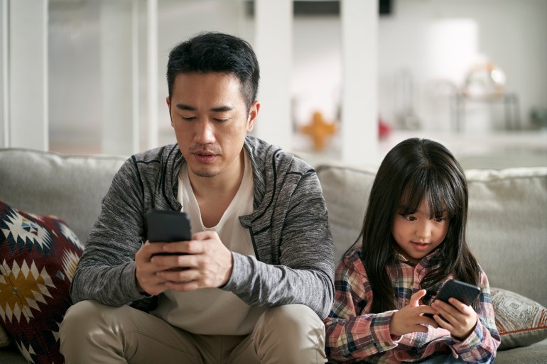 young asian father and seven-year-old daughter sitting on family couch using cellphone together looking serious شترستوك