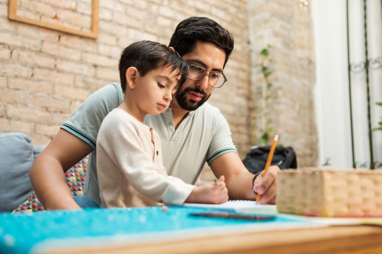 a young mexican father and his little boy sitting in front of the coffee table at home and drawing on a notebook with a pencil and a crayon.