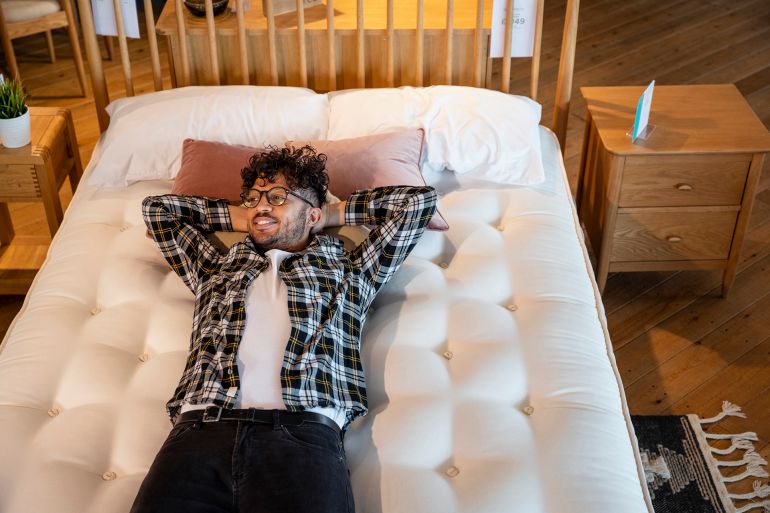 a mid adult man lying on a bed in a furniture store, he is testing it out and considering to buy it. he has his arms behind his head and is looking away from the camera.