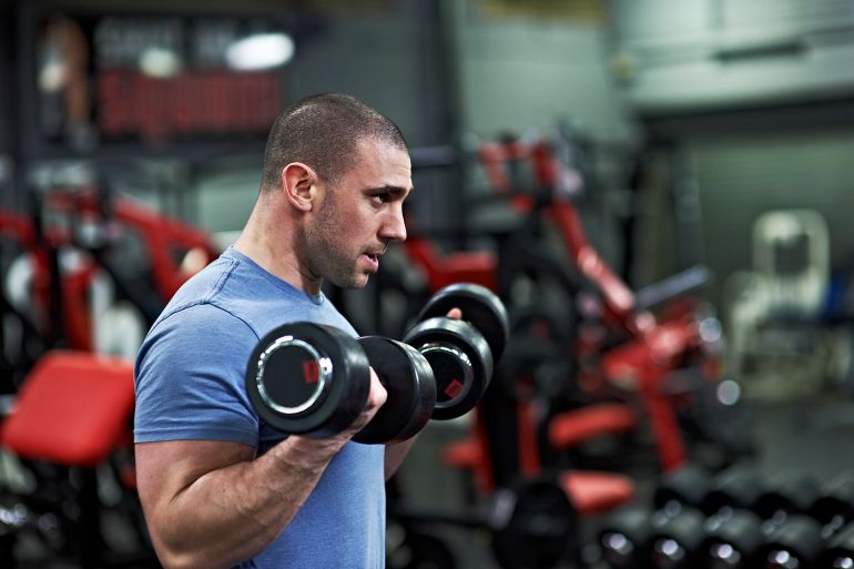 Physically strong male lifting heavy dumbbells in gym, surrounded by exercise apparatus