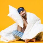 Full length of smiling young bearded man in pajamas home wear sleep mask sitting with pillow blanket isolated on bright yellow colour background studio portrait. Relax good mood lifestyle concept; Shutterstock ID 1885457752; purchase_order:aja; job:; client:; other: