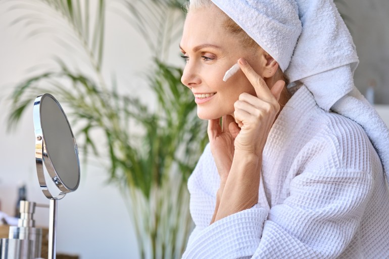 Gorgeous senior older adult 50 years old blonde woman wearing bathrobe and turban towel in bathroom applying moisturizing tightening face skin treatment, looking at mirror. Morning beauty routine.