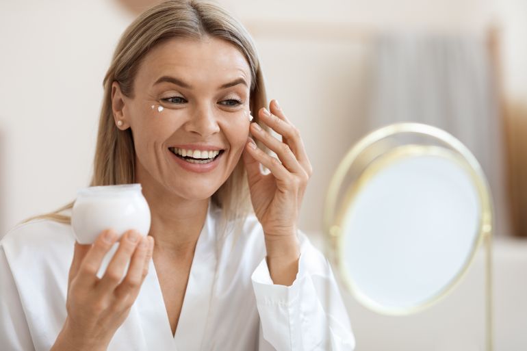 Closeup of beautiful blonde middle aged lady in silky bathrobe using nourishing eye cream, looking at mirror and ...