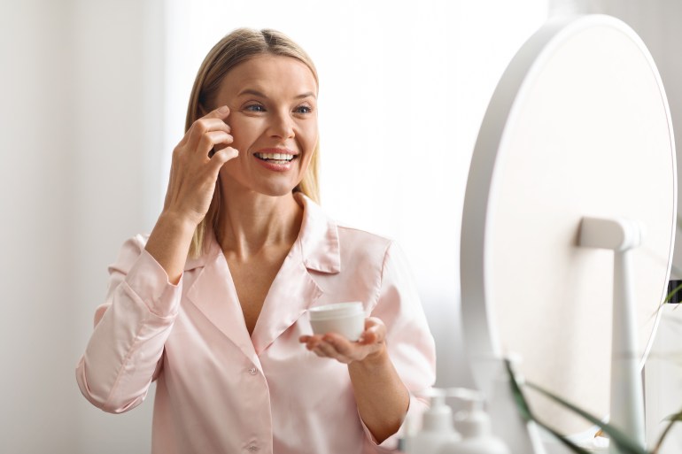 Closeup of beautiful blonde middle aged lady in silky bathrobe using nourishing eye cream, looking at mirror and ...