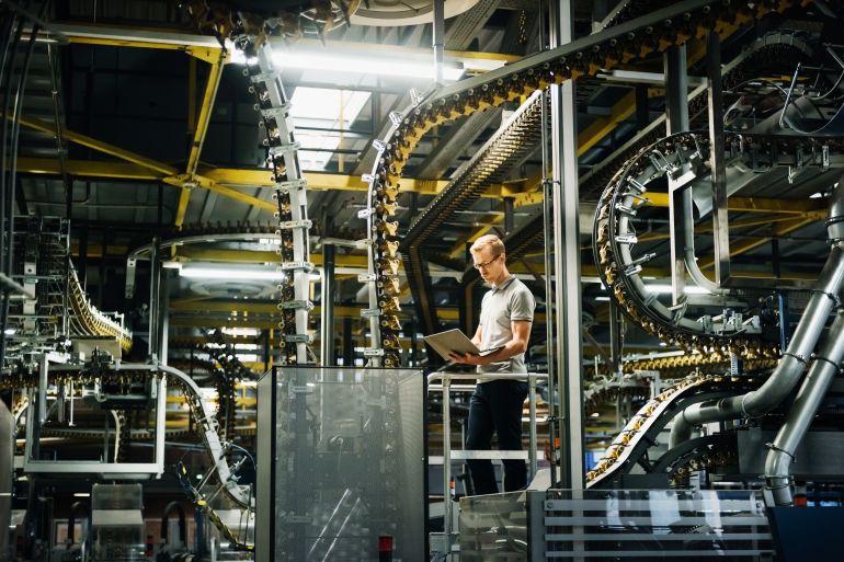 engineer holding a laptop standing in a factory between machines and conveyors