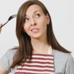 Young fun crazy brunette housewife in striped apron gray t-shirt isolated on white background Housekeeper woman holding white empty plate fork in hair like comb hairbrush. Copy space advertisement; Shutterstock ID 1026887551; purchase_order: AJA; job: ; client: ; other: