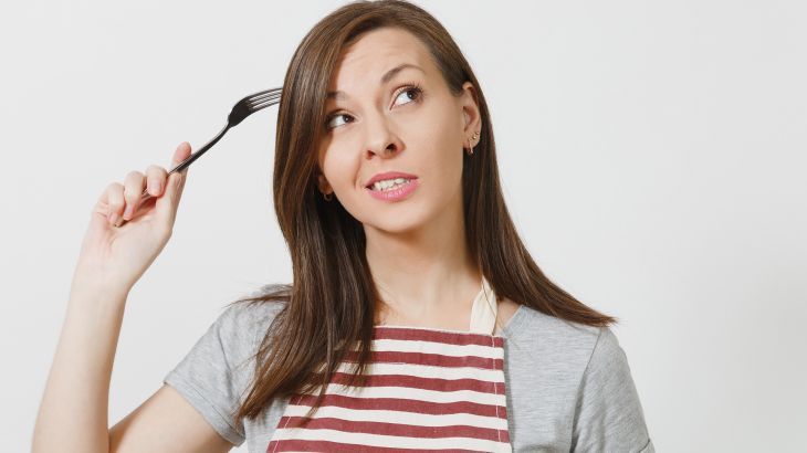 Young fun crazy brunette housewife in striped apron gray t-shirt isolated on white background Housekeeper woman holding white empty plate fork in hair like comb hairbrush. Copy space advertisement; Shutterstock ID 1026887551; purchase_order: AJA; job: ; client: ; other: