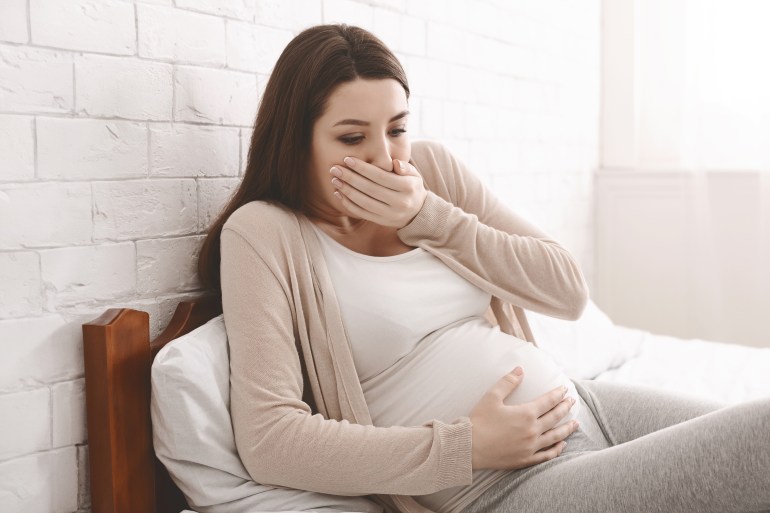 morning sickness. young pregnant woman sitting on bed, covering her mouth feeling nauseous during pregnancy, free space