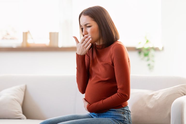 pregnancy morning sickness. pregnant european young lady having nausea feeling bad and sick, sitting on sofa and covering mouth whit hand at home interior. copy space